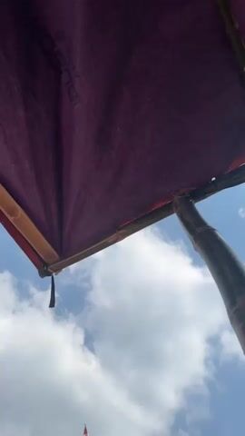 The video opens with a close-up of a purple canopy, revealing a glimpse of bamboo poles and a blue sky with white clouds. The camera then pans down, showing a tall bamboo pole adorned with numerous Indonesian flags (red and white stripes) reaching high into the sky. Several other similar flagpoles are visible in the background, along with lush green trees and foliage. In the mid-ground, two men are engaged in an activity around the base of the main flagpole. One man, wearing a blue shirt and dark pants, is watering the base of the pole with a hose, creating a spray of water. Another man, wearing a light green shirt and dark pants, is standing near the pole. A large blue barrel is positioned next to the main flagpole. A small, rustic building with a tiled roof is visible behind the men and flagpoles. The ground is covered with reddish-brown soil, and a black hose is laid across it. The overall scene depicts an outdoor setting, possibly a village or rural area, where preparations are being made, likely for a flag-raising ceremony or a celebration.