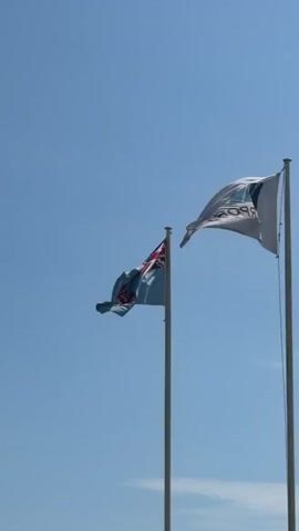 The video shows two flags on flagpoles against a clear blue sky. The flag on the left is the flag of Fiji, which is light blue with the Union Jack in the canton and a shield in the fly. The flag on the right is a white flag with a logo and the text "FIJI AIRPORTS" printed on it, which appears to be the logo of Fiji Airports. The flags are both waving in the wind. The camera is looking up at the flags.