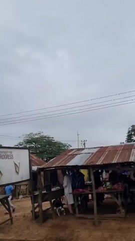 The video pans across a street market. Multiple stalls with corrugated metal roofs are visible, some displaying goods like produce and household items. Several people are walking through the market or standing at the stalls. In the background, a tall cell tower is prominent against a cloudy sky. The ground is dirt, and there are some motorcycles parked near the stalls. The overall impression is of a busy, open-air market in a developing area.