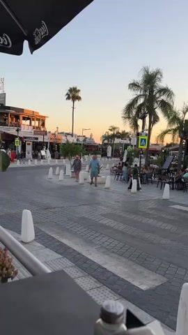The video shows a street scene at dusk. Palm trees and buildings line the street, with several outdoor seating areas for restaurants or bars. People are walking and sitting, and cars are visible in the background. The sky is clear and orange, indicating sunset. A pedestrian crossing is prominent in the foreground.