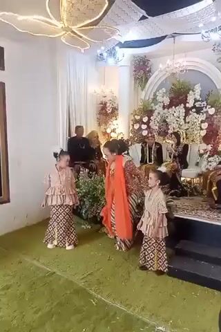 A wedding reception is in progress with a bride and groom seated behind a floral arrangement. Two young girls in traditional attire are dancing on a green carpet in front of a woman who is also dancing and directing them. The woman is wearing a red and orange outfit. The room is decorated with lights and flowers.