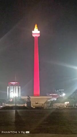 The video shows a night-time view of the National Monument (Monas) in Jakarta, Indonesia. The monument is illuminated with lights, which change color throughout the short clip. In the background, there are other buildings and city lights. The foreground shows a paved area. The image is a vertical shot and includes a watermark at the bottom left corner that reads "PHANTOM V Flip 5G".
