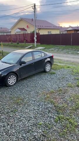 A black sedan is parked on a gravel surface in front of houses with brown fences. The sky is blue with white clouds and sunlight beams visible. A utility pole stands to the right of the vehicle.