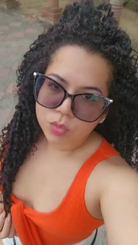 The video shows a person taking a selfie in what appears to be a public park or square. The person, who seems to be a woman, has dark curly hair, wears black-framed sunglasses, and is wearing an orange top. She poses with a pursed lips expression. In the background, there are trees, walkways, and architectural details like columns and a statue. The camera is handheld and moves slightly, indicating she is holding the phone to take the selfie. The lighting appears to be natural daylight.