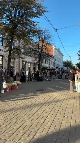 This is a vertical, medium-shot video of a street scene with people and buildings. The street is paved with brick. There are trees lining the street, with buildings on either side. In the foreground, there are people standing and observing. In the middle ground, there is a group of musicians playing instruments, possibly a band. They have audio equipment set up. Further down the street, more people are walking and some are pushing strollers. The sky is blue and the lighting suggests it is daytime, possibly late afternoon due to the long shadows. There are also some red buckets and flowers visible near the left side of the frame. The overall atmosphere appears to be lively and public.