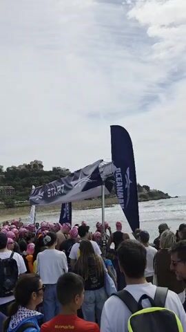 A crowd of people wearing pink swimming caps stands at the beach, gathered around a start line for a swimming race. The banner above the start line says “START” in white letters against a dark background. Behind them, the ocean extends to the horizon, and a hillside with buildings is visible. Two large blue flags with white logos stand at the sides of the crowd. People of various ages and appearances are present, some wearing backpacks and casual clothing, while others appear ready for the swim. The overall mood appears anticipatory, as the crowd awaits the start of the race. The sky is overcast, with some patches of blue visible.