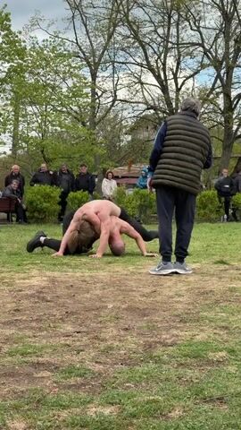 In an outdoor setting, two shirtless men wrestle on a grassy patch, their bodies intertwined in a grappling hold. They are surrounded by a group of observers, some seated on a bench and others standing. An older man with gray hair, wearing a padded vest and dark pants, stands to the right, facing the wrestlers and appearing to watch or referee the match. The background consists of trees with sparse foliage, a few buildings in the distance, and patches of dirt mixed in with the grass. The overall lighting suggests it is daytime.