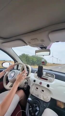 The video shows the interior of a white Fiat 500 car. A person's hands are on the steering wheel, with rings on their fingers. The dashboard is white and cream-colored. A smartphone is mounted on the dashboard. Outside the car, there are windmills in a field, and the car is on a paved road. The sky is cloudy. There is a car air freshener hanging from the rearview mirror.
