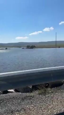 The camera is positioned behind a gray metal guardrail. A view of a body of water stretches out toward a flat, grassy area. Small islands or rock formations are scattered within the water. A transmission tower is visible in the distance. The sky is blue with scattered white clouds.  The water is rippled and reflects sunlight. The landscape appears flat and open. It's a bright, sunny day with moderate wind.