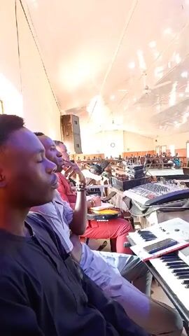 This is a first-person perspective video taken inside a large room, likely a hall or a church, with a high ceiling and numerous fluorescent lights. In the foreground, several individuals are seated, with a keyboard prominently displayed in the lower right corner. To the left, a person in a dark outfit is looking to the left. Next to them, another person in a light-colored shirt is also looking to the left, with their arms crossed. Behind them, a person in a red shirt is visible. In the mid-ground, various musical instruments and equipment are set up, including another keyboard, a sound mixing board, and speakers. In the background, the room extends further, with rows of chairs and tables, and more people are sparsely seated. The overall atmosphere suggests a gathering or a rehearsal for a musical event.
