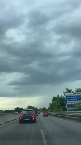 The video shows a view from inside a vehicle traveling on a highway under a cloudy sky. Several cars are visible on the road ahead, moving in the same direction. The car in the foreground is dark gray, with its taillights illuminated. A red car is further ahead in the right lane. Trees line the right side of the highway, and a concrete barrier is on the left. The clouds are dark and appear heavy, suggesting a possibility of rain in the distance. The overall atmosphere is overcast.