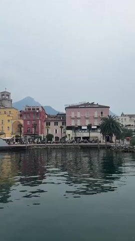 The video shows a waterfront scene in front of a row of colorful buildings. The buildings are multi-story with balconies and awnings. Some of the buildings have signs that say "HOTEL". There are palm trees and other greenery in front of the buildings. The water in the foreground is dark and choppy, reflecting the buildings and the sky. The sky is overcast. In the background, there are mountains. The camera is moving slowly to the right, panning across the scene. There are people visible on the waterfront, sitting at tables outside some of the buildings. There is also a white structure that looks like a boat or a modern sculpture in the water.