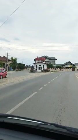 The video shows a view from inside a vehicle moving along a paved road. The road has white dashed lines marking the lanes. On the right side of the road, there is a small white building with a red-domed roof, which appears to be a church. There are rose bushes in front of the church. Across the road, on the left, there are houses and trees. The sky is overcast. The video is shot from a low angle, suggesting the camera is mounted on the dashboard or windshield of the vehicle.