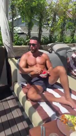 A man lies shirtless on a cushioned outdoor lounge eating fruit, wearing sunglasses and a towel. The view pans out to show other people lounging on beds with canopies and others sunbathing by a swimming pool with umbrellas.