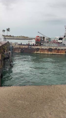 The video shows a boat or barge tied to a dock or pier. The vessel appears to be rusty and weathered. There are several people on board. Some smoke is visible in the background, near the dock. The water is somewhat choppy. The sky is overcast.  The presence of palm trees suggests a tropical or subtropical environment.