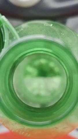 The video shows a close-up, interior view of a green plastic bottle. The focus shifts slightly throughout the recording, but the overall perspective remains consistent. The bottle's opening is visible, with the green plastic forming a circular rim. Inside, there are concentric rings and indentations, suggesting the bottle's construction. The interior is illuminated, and the color of the plastic casts a greenish hue on everything within the frame. No people or other objects are clearly discernible within the bottle. The background is out of focus and appears to be a light-colored surface with some darker elements.