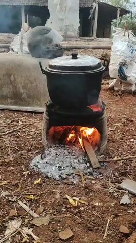 A black pot sits on a rustic, makeshift stove made of metal. Flames are visible beneath the pot, indicating it is being heated. The stove is surrounded by dirt and debris, with dry leaves and twigs scattered around. In the background, there is a dilapidated structure, possibly a house or shed, with broken walls and a partially visible roof. A black helmet is placed on a curved, concrete-like object to the left of the stove. The overall scene suggests an outdoor cooking setup in a rural or less-developed area.
