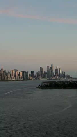 A wide shot captures a hazy, panoramic view of a city skyline across a wide body of dark water under a pale, dusky sky. The skyline is dominated by numerous skyscrapers of varying heights and architectural styles, illuminated by soft, warm light. The water is choppy, with gentle ripples and faint, circular wave patterns visible. In the foreground, a dark, irregular landmass with a few small buildings and trees juts out into the water from the right side of the frame. The overall atmosphere is calm and serene, with the fading light casting a peaceful glow over the urban landscape.