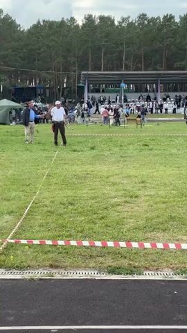 The video shows an outdoor event with spectators seated in bleachers in the background, under a canopy. The foreground is a grassy field with several people scattered about. In the middle distance, there are tents set up and more people gathered. The sky is cloudy. The video pans from left to right, showing different areas of the event.