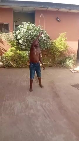 A barefoot, shirtless, dark-skinned youth in denim shorts manipulates a bow and arrow on a concrete surface in front of a two-story peach colored building. The youth appears to be pulling back on the bow and aiming. The background includes green bushes and plants against the wall of the building, a window, and a light fixture.  The scene appears to be outdoors in daylight. There is indistinct background noise.