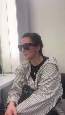 A woman is sitting in front of a mirror wearing oversized sunglasses and a striped shirt over a black t-shirt. She has her hair pulled back and is wearing a gold cross necklace. She makes a face with pursed lips and then smiles slightly. The background is a plain white wall with a vertical panel to the left that reflects green light. A white countertop is partially visible in the foreground. She is wearing a black purse over her shoulder.