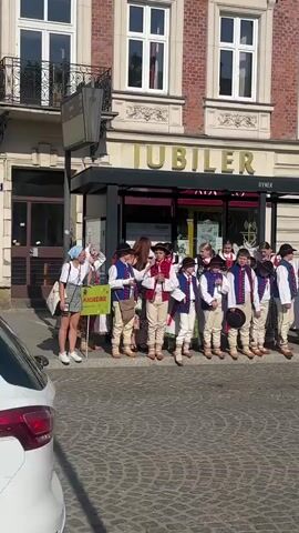 A group of children in traditional Polish folk costumes stand in a line on a cobblestone street. They are in front of a building with a sign that reads "JUBILER." The children are wearing white shirts, red vests, and beige pants. Some of them are wearing hats. A woman is standing to the left of the children, holding a green sign. A silver car is parked on the right side of the frame. The sun is shining, casting shadows on the street.