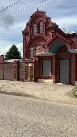 The video shows a medium shot of a brick building, from across a paved road. The building is red brick, with an ornate upper story and arched windows. The ground floor has multiple doors and windows, which appear to be boarded or painted over. The building is adjacent to a brick fence and a patch of overgrown weeds. Power lines run overhead. The sky is cloudy with patches of blue. The camera is stable and there are no apparent people.