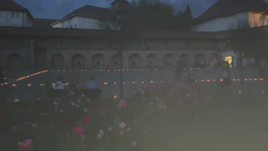 The video shows a group of people gathered in a courtyard at dusk. The courtyard is decorated with rows of candles, which create a warm and inviting atmosphere. There are buildings in the background with arched walkways. Some people are standing and talking, while others are sitting on benches. There are also some flowers in the foreground.