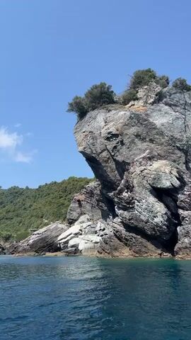 The video shows a body of water with a large rock formation on the right side of the frame. The rock is tall and jagged, with trees growing on top. To the left of the rock formation is a green, forested hillside. The sky is blue and clear. The water is a deep blue color and appears to be calm.