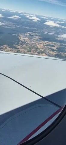 The video shows a view from an airplane window looking down at the ground and out at the sky. The airplane wing is visible in the foreground, taking up the bottom half of the frame. The wing is light gray and has a dark line running diagonally across it. The ground below is a patchwork of green forests, brown and tan fields, and scattered buildings. There are also some bodies of water visible. The sky is blue with scattered white clouds. The camera is moving slightly, suggesting the airplane is in flight.
