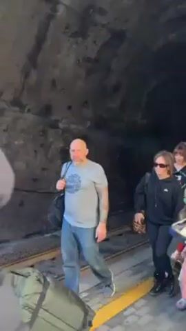 Several people are walking in a dark tunnel. The tunnel has a visible track in the middle with a narrow walkway on each side. The group comprises women, men, and children of varying ages. The people are walking with purpose, seemingly heading towards the tunnel's exit. The adults are wearing casual clothing, while the children wear a variety of colorful tops. The light within the tunnel is limited, highlighting the textures of the rocky walls.