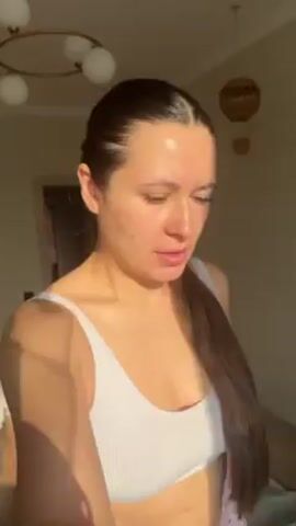 A woman with long, dark hair tied in a sleek ponytail is shown in a sunlit indoor setting. She is wearing a white ribbed sports bra or crop top, and brief glimpses of light pink patterned shorts are visible at the end of the clip. The bright sunlight streams in from the right side, casting soft shadows across her face and body, and highlighting her features.

She looks directly at the camera with a warm, friendly smile, then shifts her gaze downward, maintaining a pleasant and content expression. Her movements are subtle; she slightly adjusts her posture and looks around briefly. The woman appears to be in a relaxed and happy mood. In the background, a modern ceiling light fixture with multiple globes is visible on the left, and a woven wall decoration hangs on the right. The walls are a light, neutral color.