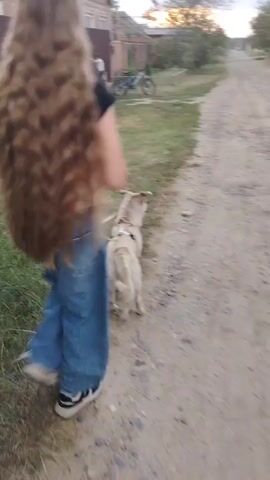 A person with very long brown hair is walking down a dirt road, possibly with a dog on a leash. The person is wearing loose-fitting blue jeans and a dark t-shirt. There are houses and trees on either side of the road, and the sky is cloudy with a hint of sunset. A bicycle is parked near one of the houses.