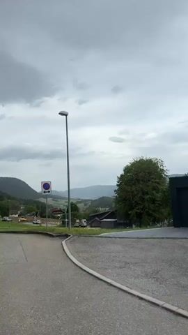 The video shows a view of a rural landscape under a cloudy sky. In the foreground, there is a paved area with a grassy edge. Several trees are visible, some with thick foliage. In the middle ground, there are buildings, including what appears to be a municipal building with the word "MUNICIPALITE" visible, and other houses or structures. Cars are parked in a lot. Beyond the buildings, rolling hills covered in greenery extend into the distance, with more hills or mountains forming the backdrop. The overall impression is of a quiet, somewhat overcast day in a village or small town surrounded by nature.