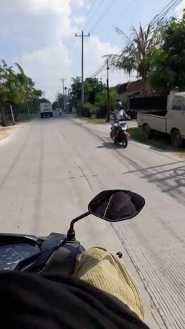 The video is shot from the perspective of someone riding a motorcycle. The rider's hand is visible, wearing a light-colored glove. The motorcycle's rearview mirror is also in view, reflecting a person's face and upper body. The road ahead is paved with some gravel on the side. On the right side of the road, there are trees, houses, and a small mosque with a red-tiled roof. On the left side of the road, there are more trees, houses, and a white truck. The sky is cloudy. The video appears to be taken during the daytime.