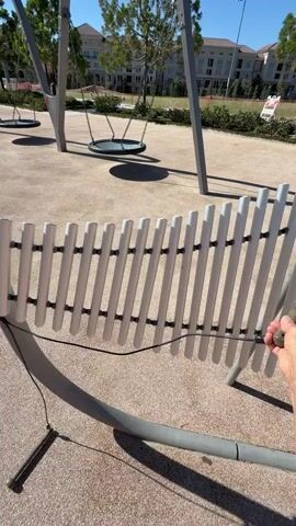 A person's arm and hand are visible playing a set of outdoor chimes. The chimes are made of white metal tubes of varying lengths arranged in a curve. A mallet, attached by a cord, is being used to strike the chimes. In the background, a playground with swings is visible, along with buildings and trees under a clear sky. The ground surface is a tan, sandy material.