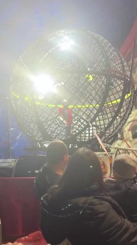 A static eye-level shot captures three people watching a motorcycle stunt inside a large, spherical cage. The cage is made of intersecting metal bars and has a bright light inside. One motorcycle rider, dressed in red, rides along the interior of the sphere. The three audience members are seen from the back. The person on the left is a man with short dark hair, wearing a black jacket. The middle person is a woman with long dark hair, also in a black jacket. The person on the right is wearing a grey hat. They are seated in front of the cage, with glimpses of staging and circus-themed backdrops surrounding the cage. The lighting is predominantly artificial, creating a stage-like atmosphere.