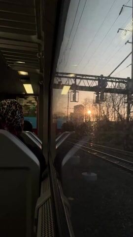 The video shows a view from inside a moving train. The camera is positioned looking out a window. A person with a leopard print head covering is seated facing forward, partially visible. The window is slightly dirty. Outside, the setting sun shines brightly, creating glare on the window. The view shows a railway track and the surrounding landscape, including trees and utility poles. The train appears to be moving relatively quickly, as suggested by the changing scenery.