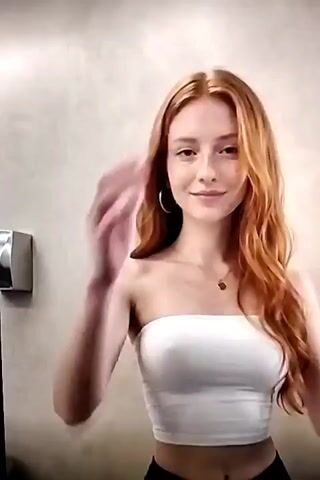A young woman with long, wavy red hair is standing in front of a light brown wall. She is wearing a white tube top and black pants. Her hands are resting on her hips and she is looking at the camera. There is a silver object on the wall to her left, possibly a hand dryer. The lighting is soft and even.