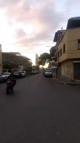 The video shows a street scene with several cars parked on the side and moving in the distance. Buildings are visible on both sides of the street. The sky is overcast with some hints of sunlight, suggesting it might be dawn or dusk. Two people are visible on the street near a white truck in the foreground.