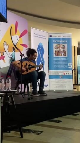 This media file captures a live performance by a musician playing an oud, a short-neck lute-like instrument. The musician, a person with dark, curly hair and glasses, is seated on a black chair on a stage, deeply engrossed in their playing. They are wearing a blue t-shirt and blue jeans, with dark shoes. The oud has a pear-shaped body and a fretless neck, and the musician's hands are actively engaged in strumming and fingering the strings.

The stage is set against a backdrop of several large banners. To the left, a colorful banner features a stylized, cartoonish illustration of a yellow character with its arms raised and pink elements resembling petals or flames. To the center, a white banner with a blue ribbon graphic displays Romanian text that reads, "Pentru o lume în care refugiații sunt bineveniți" (For a world where refugees are welcome) and "#WithRefugees." To the right, a larger blue banner titled "INTEGRIS" features an illustration of a diverse group of people holding hands around a tree, with Romanian text below. This banner also displays the European Union logo and other official seals.

In the foreground, to the left of the stage, is a small black table with two bottles of water and what appears to be a microphone stand. The stage floor is black. The overall setting suggests an indoor event, possibly a cultural festival or an awareness campaign related to refugees, given the text on the banners. The lighting is consistent with an indoor venue, and the time of day cannot be determined. The musician appears focused and dedicated to their performance. The overall mood is one of artistic expression and a commitment to a cause.