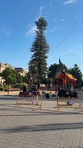 The video shows a public square. In the foreground, there are yellow safety barriers. Behind the barriers, a young person is standing near a tall evergreen tree that has been decorated with a metal structure, possibly for a festival or event. To the left, a building with an arched entrance and multiple windows is visible. In the background, to the right, a market stall with an orange canopy is set up, displaying various items for sale. The sky is clear and blue with a few white clouds. The ground is paved with interlocking bricks. The overall atmosphere appears to be that of a lively outdoor gathering.