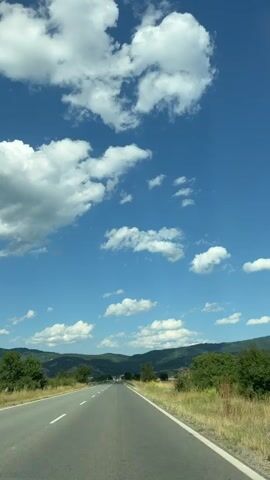 This media file captures a scenic view of a rural road in Yahinovo, Bulgaria, under a bright blue sky dotted with fluffy white clouds. The perspective is from a vehicle traveling along a paved road, indicated by the white lane markers and the guardrail visible on the right side. The road stretches towards a distant landscape of rolling green hills, some covered with vegetation and others appearing as cultivated fields, likely agricultural land.

The time of day appears to be daytime, likely late morning or early afternoon, given the clear sky and the quality of light. The weather is fair and sunny, with the scattered clouds suggesting a pleasant day. The overall mood conveyed is one of peaceful travel and appreciation for the natural beauty of the Bulgarian countryside. There is no visible text in the media file. The scene is devoid of people, focusing entirely on the landscape and the journey. The activity is simply traveling along the road, observing the surroundings. The presence of trees and dry grass along the roadside further anchors the setting in a rural, possibly summer or late spring environment.