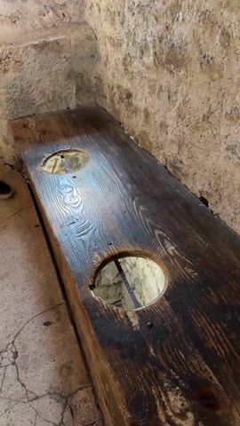 The video shows a close-up of an old wooden bench with two circular holes cut into it. The wood is dark and appears aged, with visible grain and imperfections. Light shines through the holes, revealing a stone structure beneath. The bench is positioned against a rough stone wall and the floor appears to be cracked and aged stone as well. The camera zooms in to focus on one of the holes.