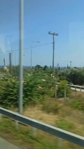 The video shows a view from a moving vehicle, likely a car or bus, on a sunny day in Greece. The scene is a coastal area with a clear blue sky and sparse vegetation, including dry grass, bushes, and trees. In the distance, the sea is visible, suggesting a Mediterranean setting.

The primary activity is travel along a road that curves through the landscape. There are road signs, streetlights, and barriers along the roadside, indicating a developed infrastructure. A cell tower is visible in the early frames, and a distant white car and a truck are seen on another road in the distance. The camera perspective is from inside the vehicle, looking outwards. Reflections on the window are also visible.

The time of day appears to be late morning or early afternoon, given the bright sunlight and the shadows cast. The weather is clear and sunny, with no clouds in the sky. The location is identified as Plaka, Litohoro, Greece, which aligns with the coastal scenery and the bright, Mediterranean atmosphere. No people are clearly identifiable, but the presence of the vehicle implies occupants. The overall mood is one of travel and observation of a pleasant, scenic environment. No significant text is clearly legible, though some road signs are present but blurred by motion.