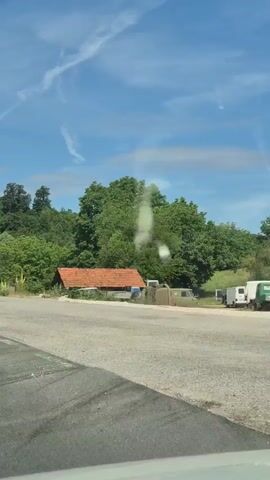 The video shows a view from a moving vehicle on a paved road. The road curves to the left. On the right side of the road, there is a metal guardrail, and beyond that, lush green trees and vegetation. In the distance, there is a small, rustic building with a red-tiled roof, partially obscured by trees and several old vehicles. The sky is blue with some wispy white clouds. There are traffic signs indicating a curve ahead. The overall scene depicts a rural or semi-rural landscape on a sunny day.