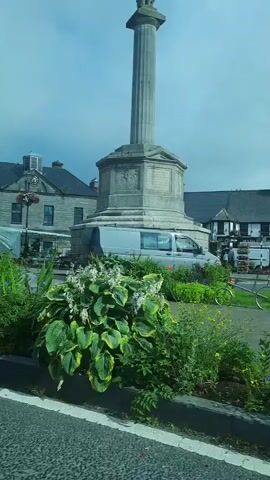 The video shows a moving view of a town square. In the foreground, there is a road and a flower bed with green foliage and some red flowers. Behind the flower bed, a large stone monument with a column stands prominently. Further back, there are buildings. One building is a light-colored stone structure with a clock tower. Another building is painted bright yellow with white trim and has awnings and outdoor seating in front of it, suggesting it is a cafe or bar. There are cars parked along the street. The sky is overcast.