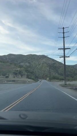 The video shows a view from inside a car driving down a two-lane road. A mountain covered with greenery is visible in the background. Power lines run parallel to the road on the right side. Several vehicles, including a red sedan, a dark pickup truck, and a white sedan, are driving on the road in the opposite direction. The sky is blue with some wispy clouds.