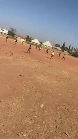 The video shows a soccer game taking place on a reddish-brown dirt field under a clear blue sky.  A group of people, presumably players, are spread across the field, most wearing brightly colored jerseys. There are buildings with white pyramidal roofs visible in the background. The camera appears to be handheld and held low to the ground, and a discarded plastic bottle lies in the foreground.  There is a tree on the left edge of the video. The players are in various positions, some running, some standing, seemingly engaged in playing soccer.