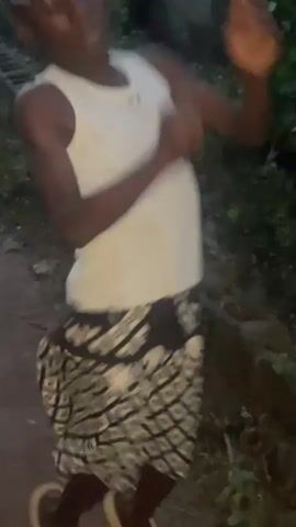 A person is dancing in a dirt path. The person is wearing a white tank top and a patterned skirt. The person is wearing white sandals. The background is a rural or village setting with buildings and vegetation. The lighting suggests it is either dusk or dawn. The camera is moving, causing some blur in the footage.
